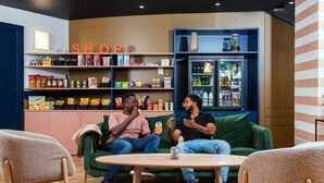 Kaksi miestä istuu vihreällä sohvalla huoneessa, jossa on hyllyjä täynnä naposteltavaa ja jääkaappi.