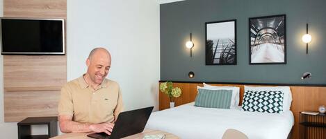 Una camera d'albergo con un letto, una scrivania con un portatile, un tavolo rotondo e una TV fissata al muro.