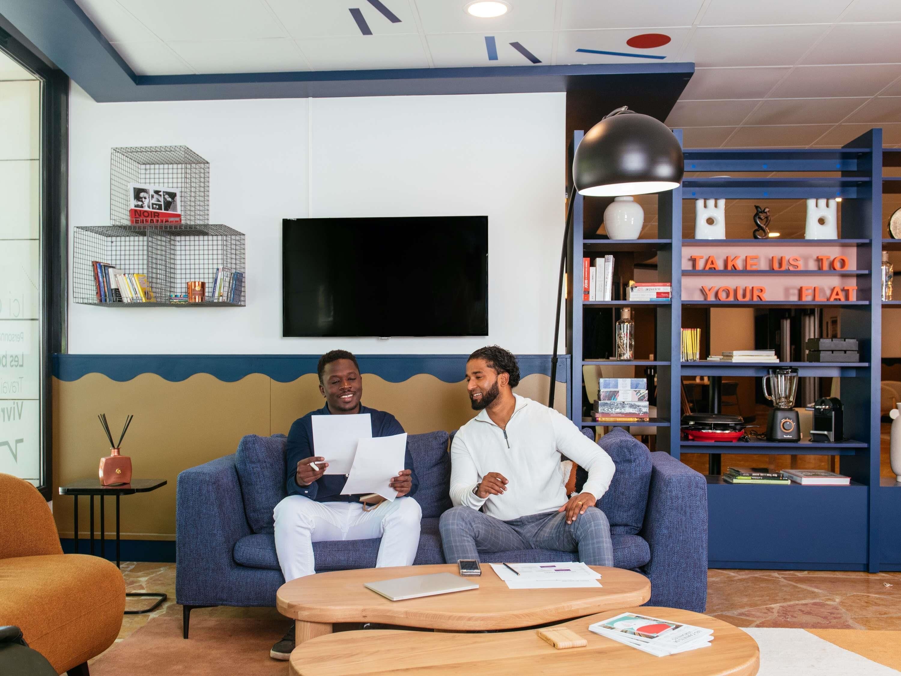 A modern lounge area with a blue sofa, a wooden coffee table, and a bookshelf with various items.