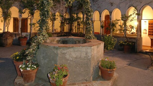 Un patio con una fuente en el centro, rodeado de plantas en macetas y un muro de piedra.