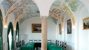 Un comedor con una larga mesa dispuesta para una comida, techos abovedados con frescos y cuadros enmarcados en las paredes.