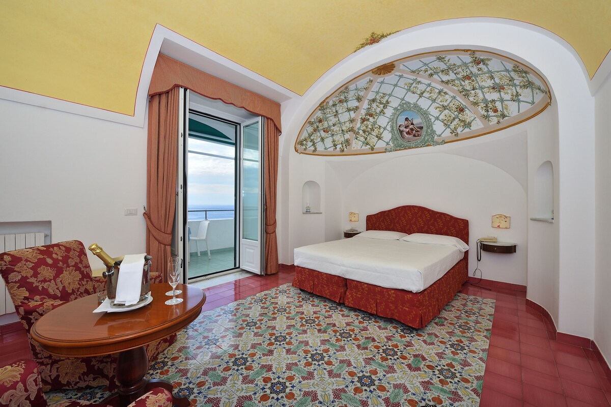 A hotel room with a bed, a table with a bottle of champagne, a chair, and a view of the sea through a glass door.