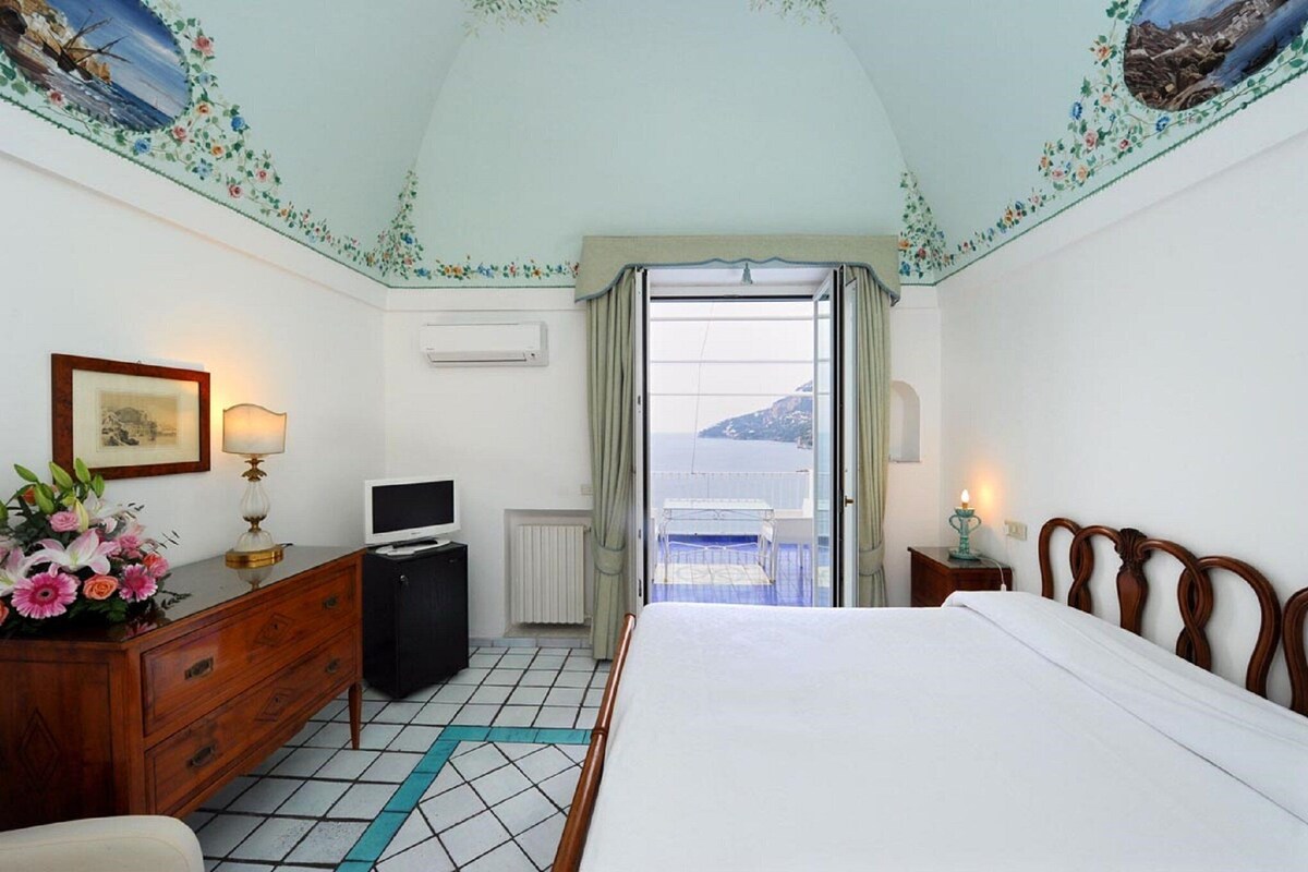 A hotel room with a bed, dresser, TV, and a view of the sea through an open door.