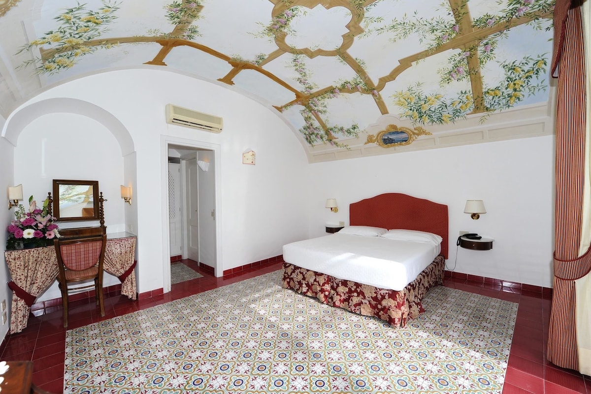 Een hotelkamer met een bed, een bureau met stoel, een spiegel en een decoratief plafond.