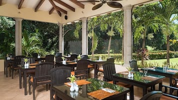 Un espacio exterior techado para comer con mesas y sillas, rodeado de abundante follaje y vistas al jardín.