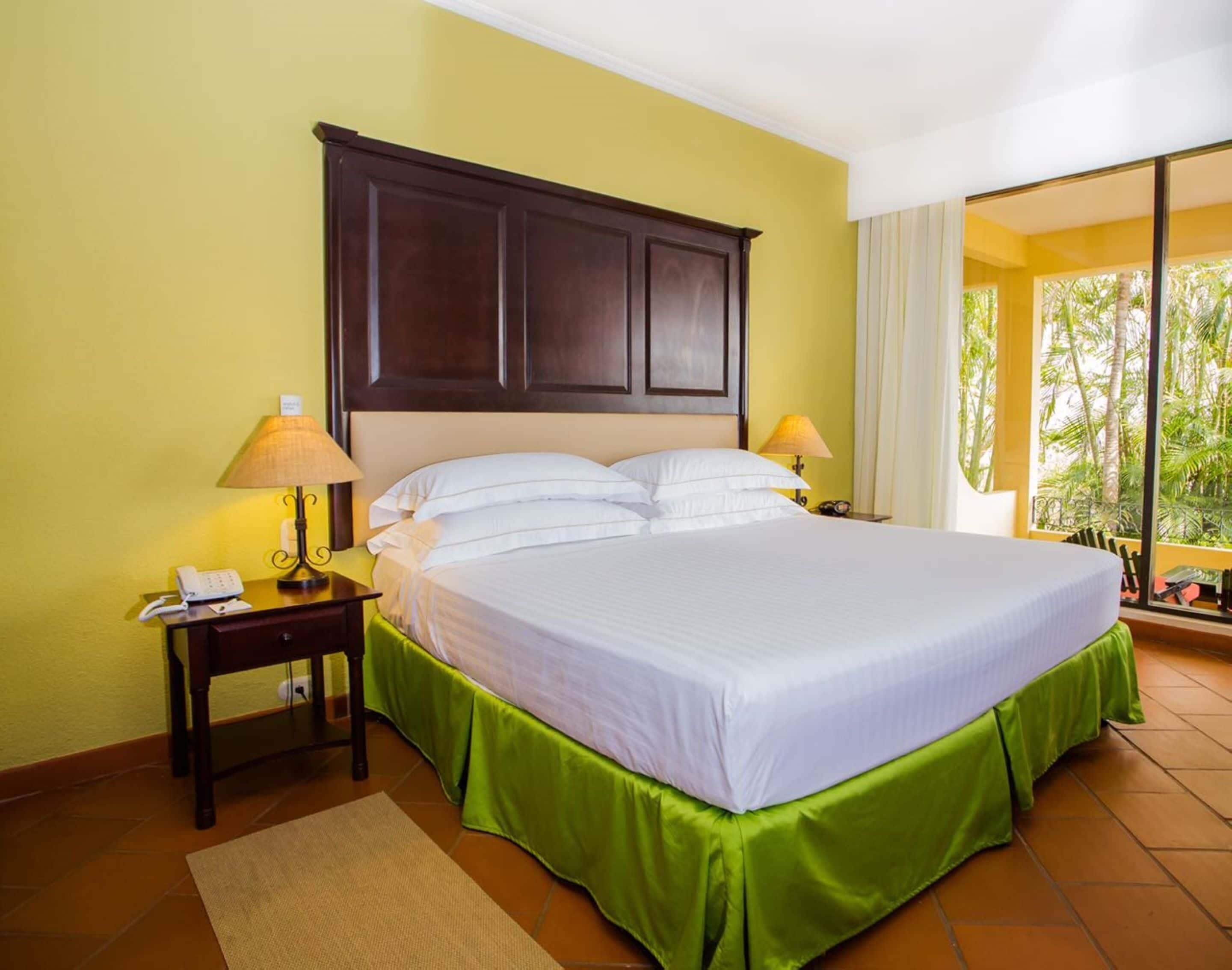 Habitación de hotel con una cama grande, dos mesitas de noche, una falda de cama verde y vistas a palmeras a través de la ventana.