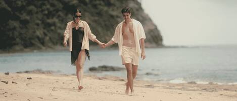 Two people walking hand in hand on a sandy beach with a rocky cliff in the background.