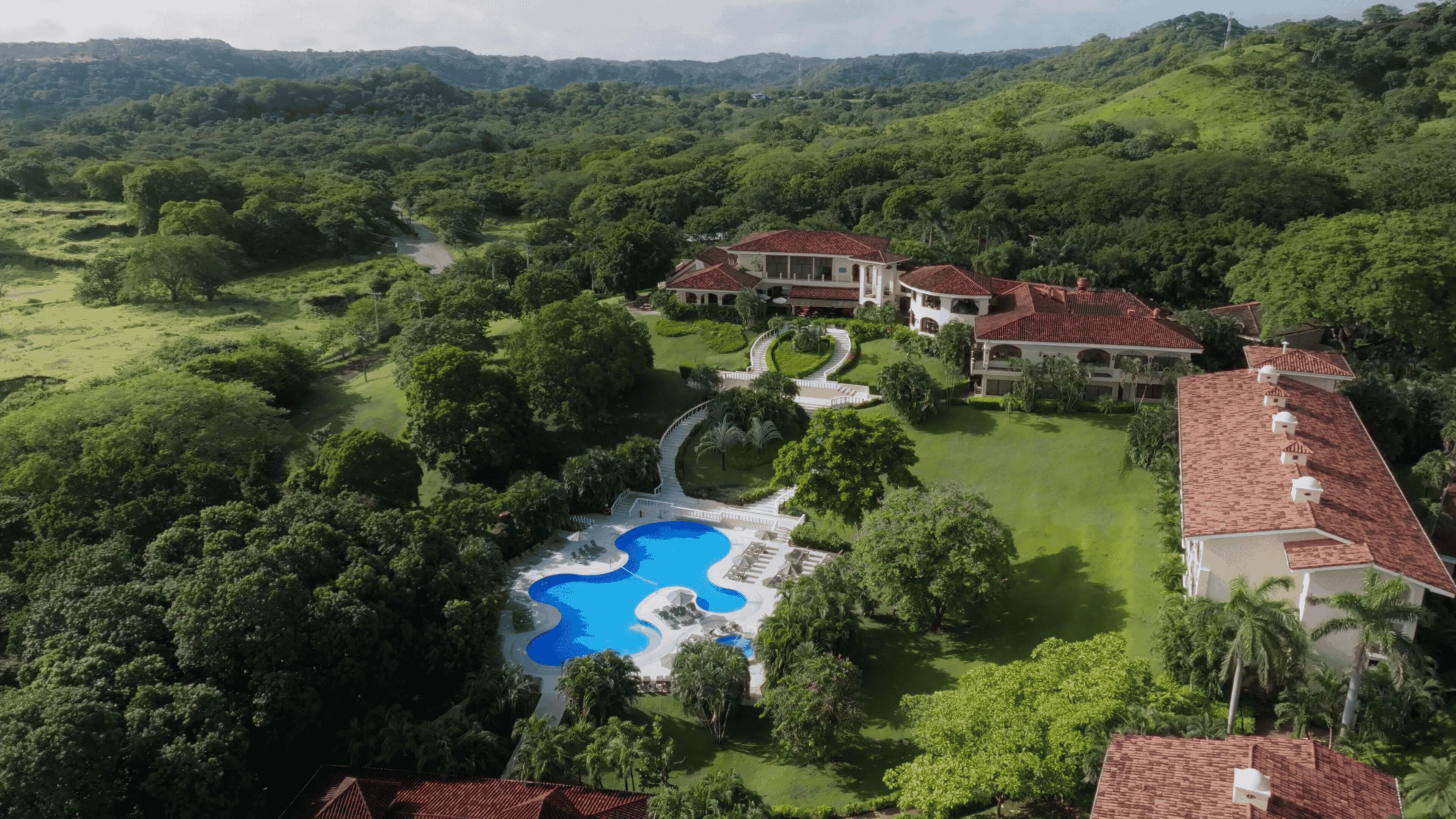 A large, luxurious estate with a swimming pool, lush greenery, and multiple buildings with red-tiled roofs.