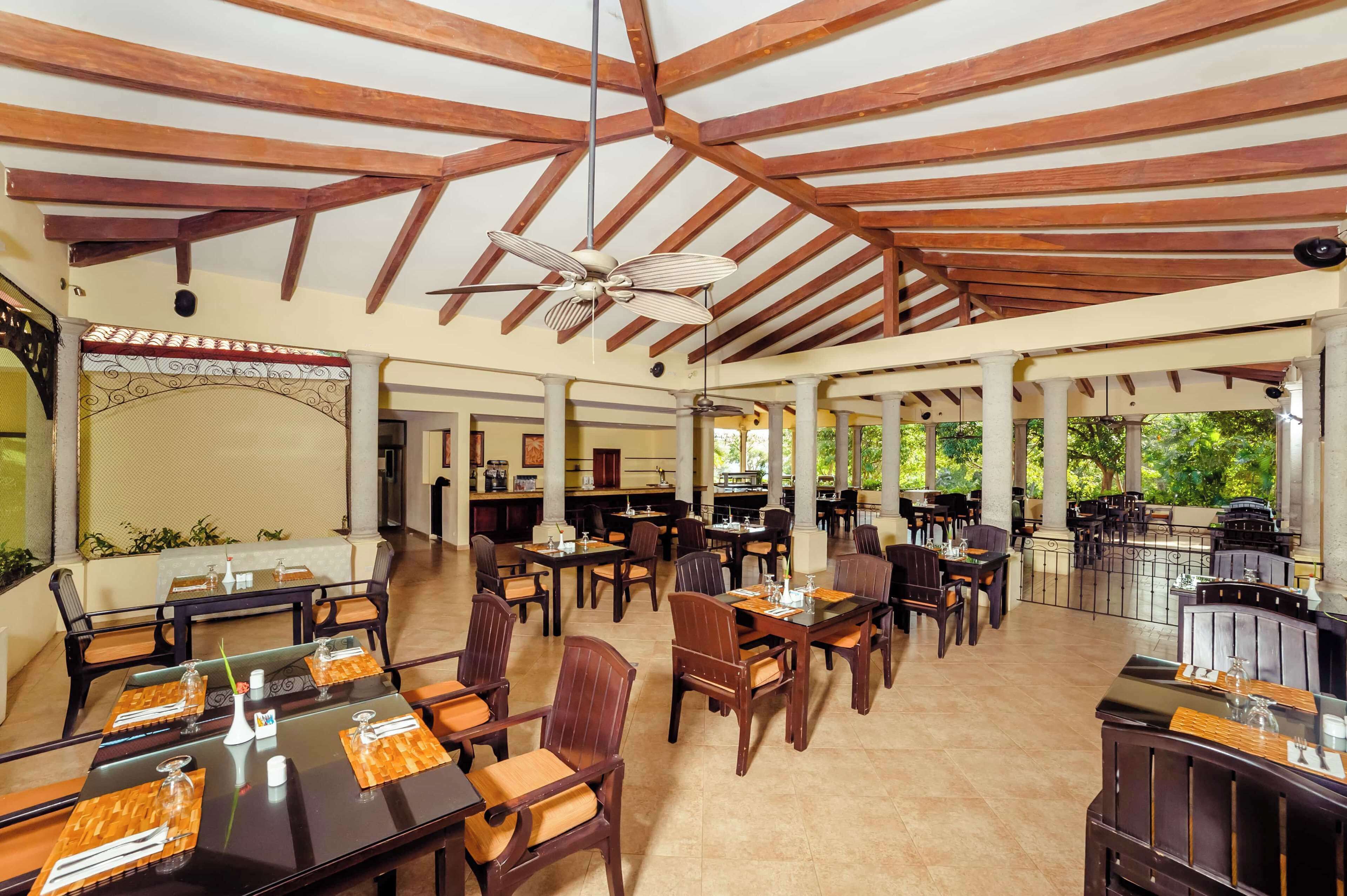 A covered outdoor dining area with wooden beams, tables with chairs, and a ceiling fan.
