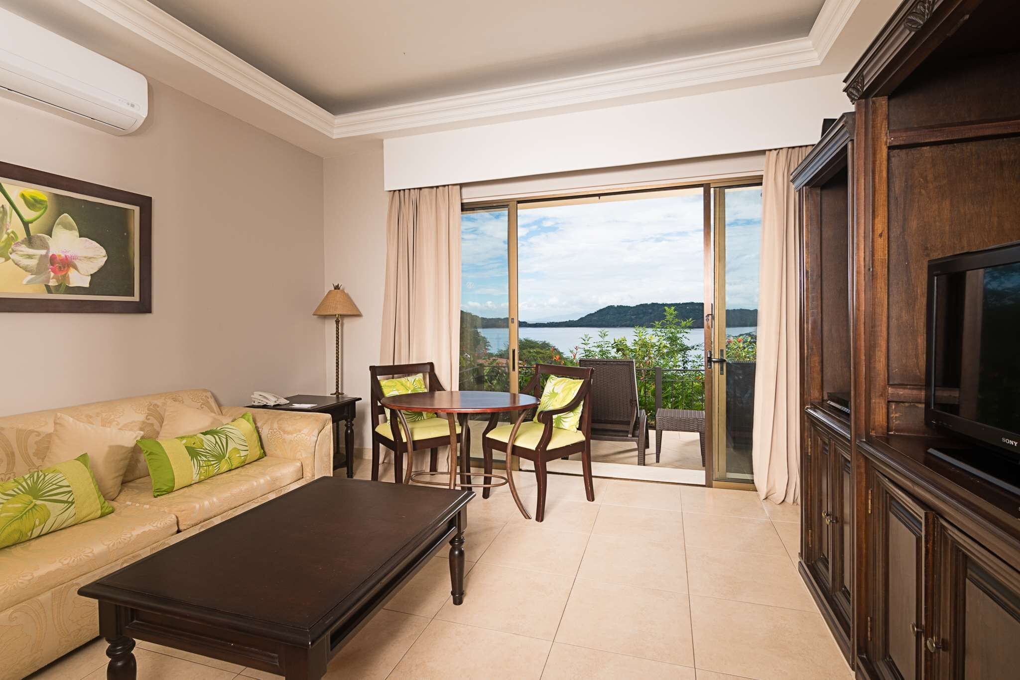 A living room with a sofa, coffee table, dining table, chairs, a TV, and a view of a lake through large glass doors.