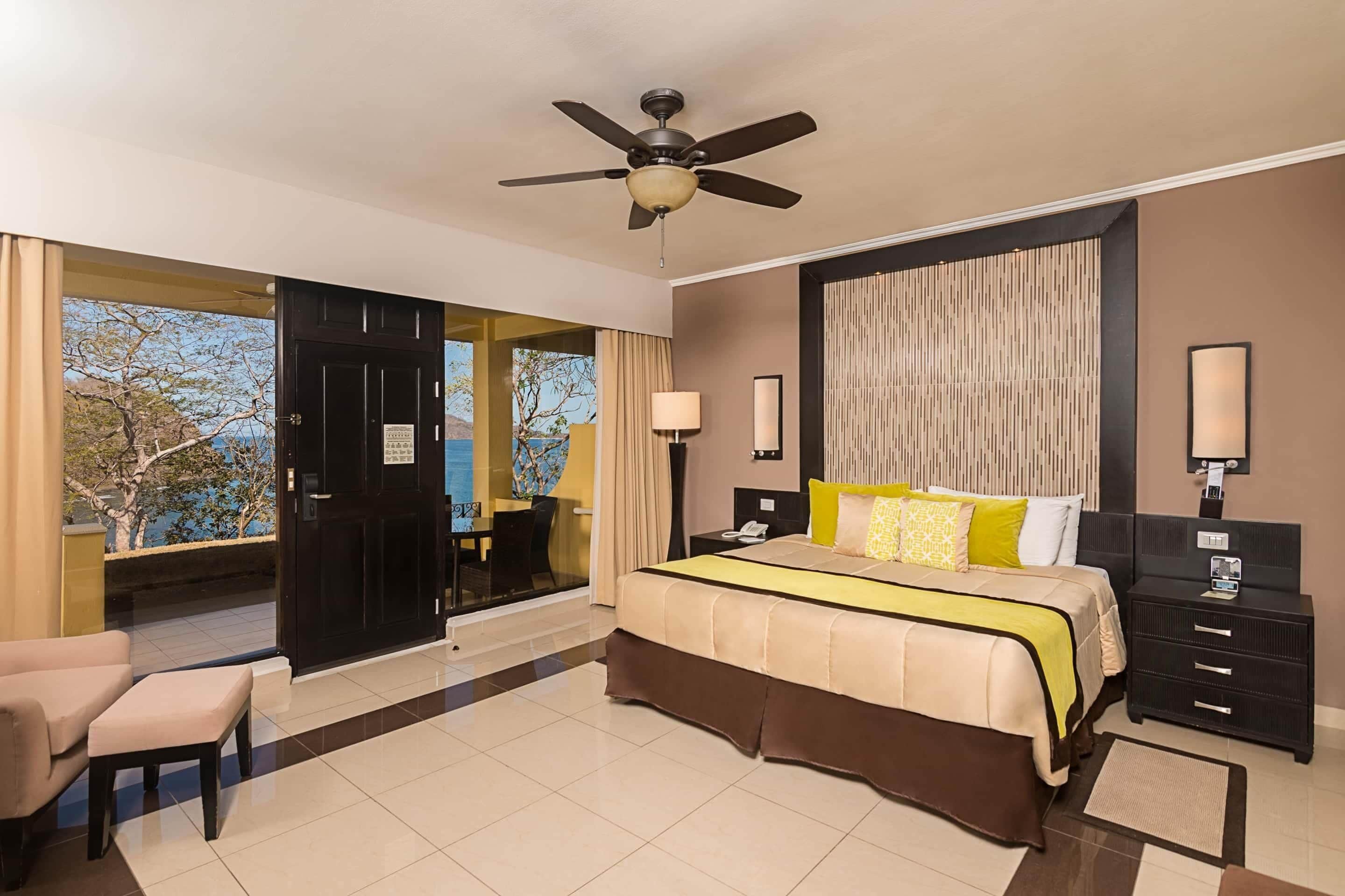 A hotel room with a bed, a desk, a chair, a ceiling fan, and a view of the outdoors.