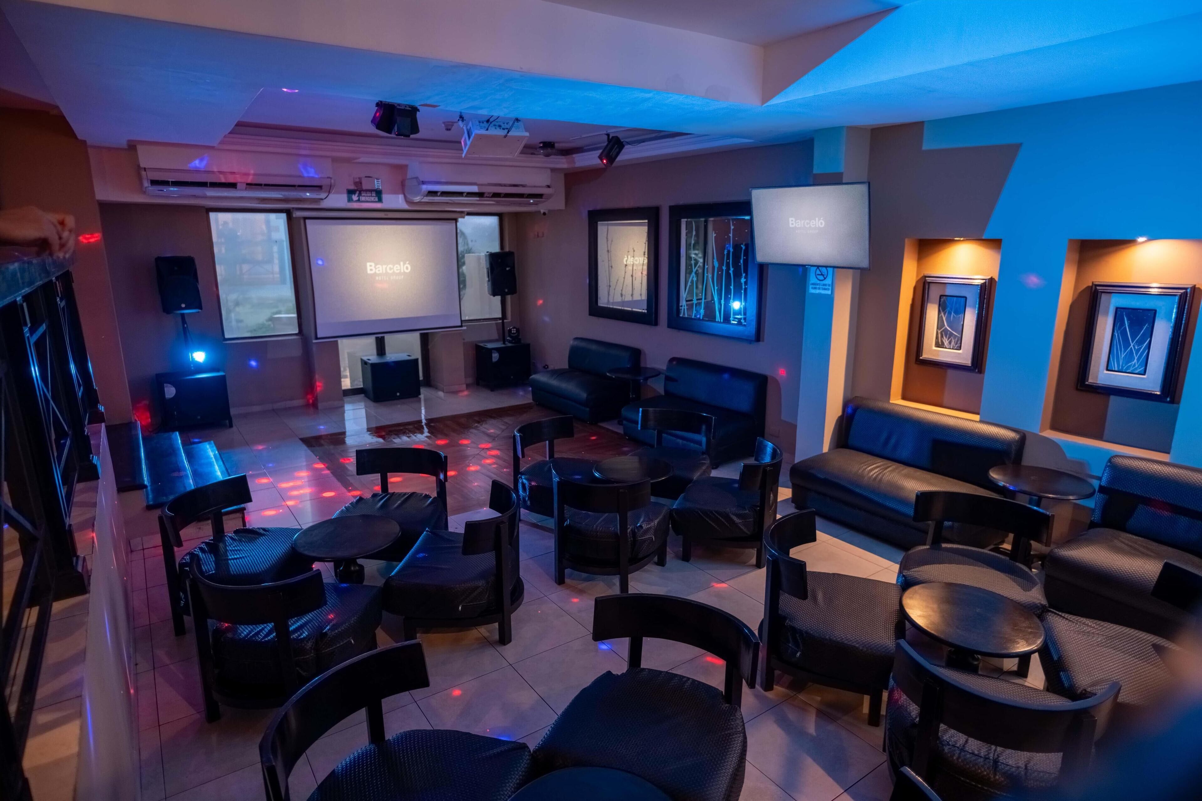 A modern lounge area with multiple seating options including sofas and chairs, a large screen displaying 'Barcelona', and ambient lighting.
