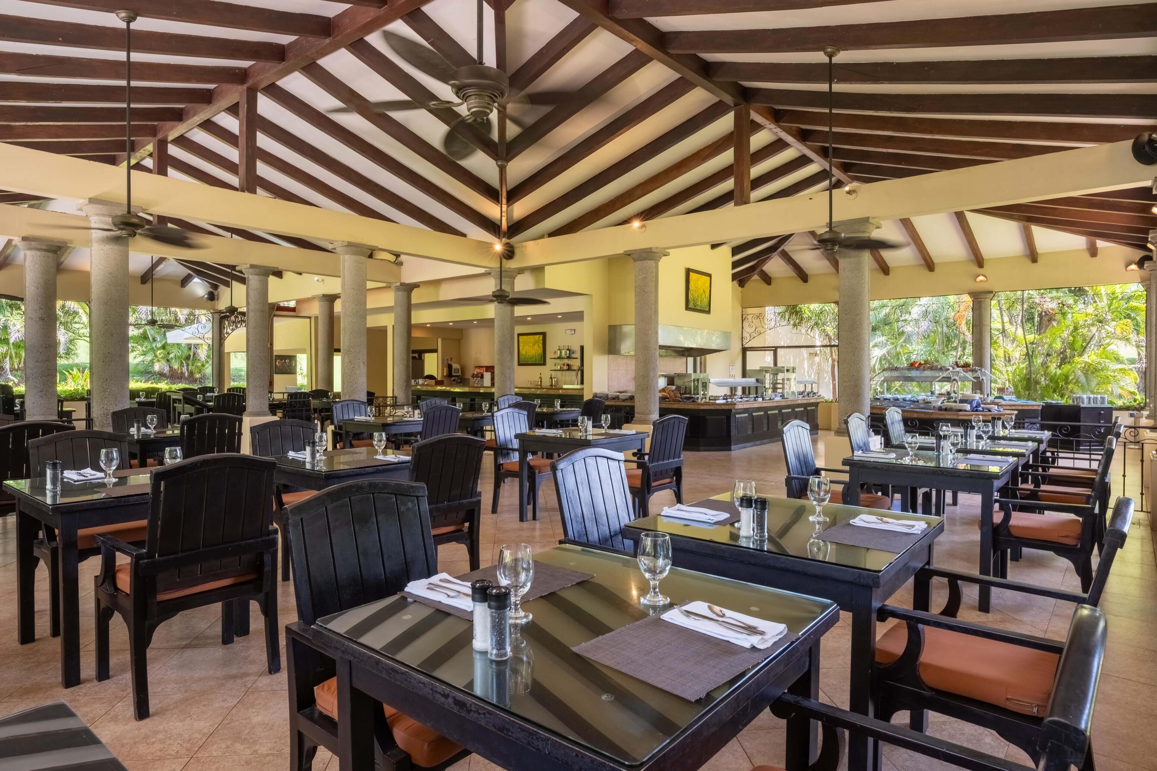 Un vaste espace extérieur aménagé pour les repas, avec plusieurs tables et chaises, un ventilateur de plafond et une vue sur la verdure environnante.