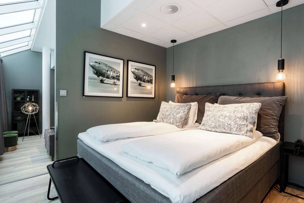 Une chambre à coucher moderne avec un grand lit, deux tableaux encadrés au mur, un banc et un bureau avec une lampe.