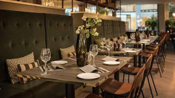 A restaurant with a long dining table set for a meal, cushioned seating, and a vase with flowers.