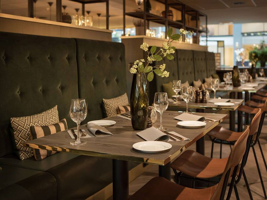 A restaurant with a long dining table set for a meal, cushioned seating, and a vase with flowers.