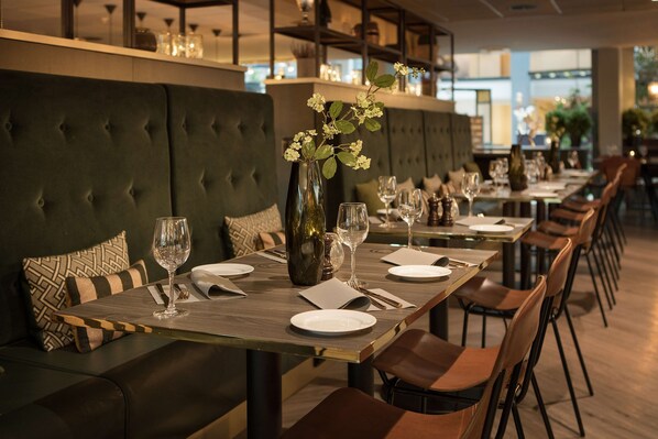 A restaurant with a long dining table set for a meal, cushioned seating, and a vase with flowers.
