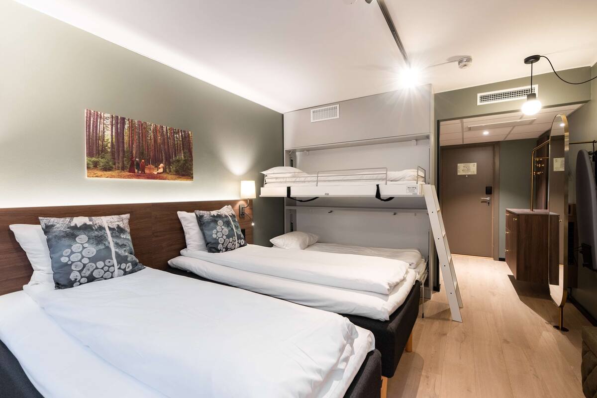 Une chambre d’hôtel avec deux lits simples, un lit superposé, un grand miroir et un tableau représentant une forêt.