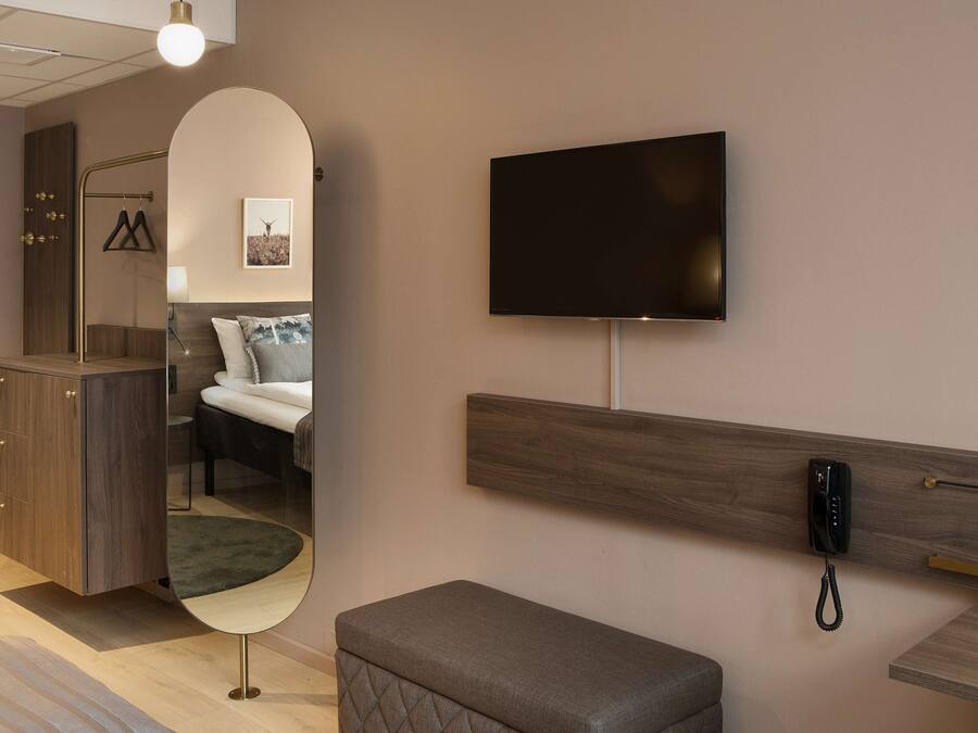 A hotel room with a bed, a bedside table, a mirror, a TV mounted on the wall, and a telephone.