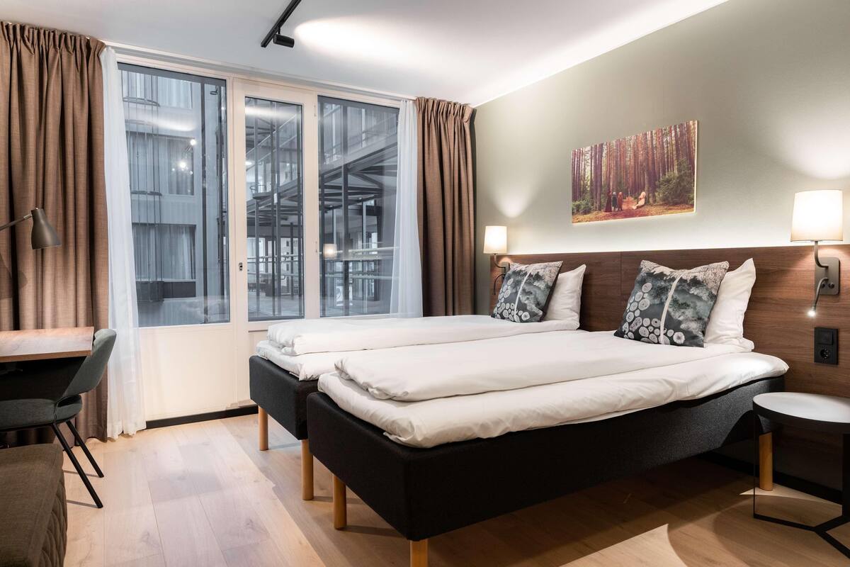 Une chambre d’hôtel moderne avec un grand lit, un bureau et une vue sur un immeuble aux portes vitrées.