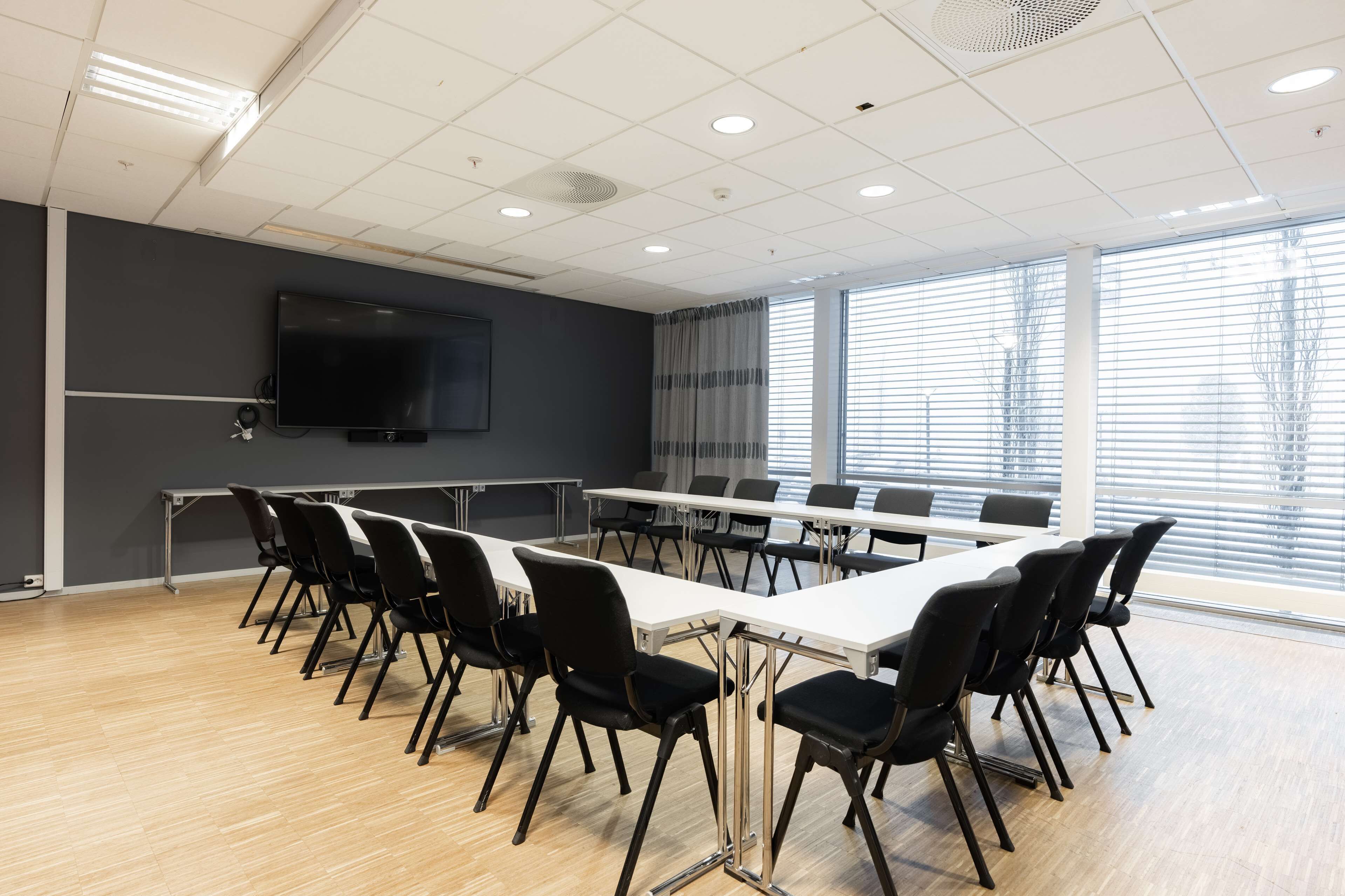 Een congreszaal met een lange tafel, zwarte stoelen, een flatscreen-tv en grote ramen met zonwering.
