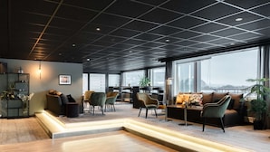 A modern lounge area with a wooden floor, multiple seating options including sofas and chairs, a coffee table, and a large window offering a view of the cityscape.