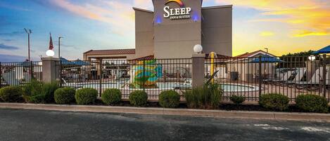 A Sleep Inn & Suites building with a sign, a fenced pool area, and a sunset sky.