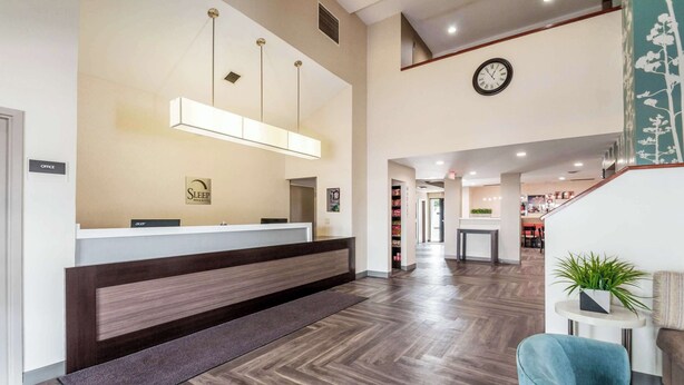 A hotel reception area with a wooden reception desk, a clock on the wall, and a staircase leading to an upper level.