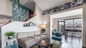 A modern living room with a sofa, two armchairs, a coffee table, and a small side table with a plant. A staircase with a green wall and a picture frame is visible in the background.