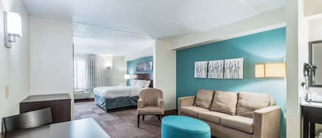 Una habitación de hotel con cama, sofá, sillón, mesa y un pouf azul.