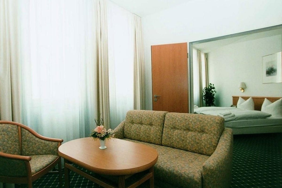 A hotel room with a sofa, a small round table with a vase of flowers, a bed with a white comforter, and a wooden door.