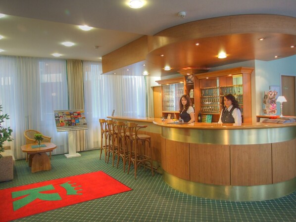 A hotel reception area with a curved counter, wooden chairs, a red carpet, and a bulletin board. - Kubrat In Helle Mitte (Berlin)