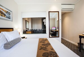 Hotelkamer met een bed, nachtkastje, lamp, spiegel en uitzicht op de badkamer en keuken door een raam.