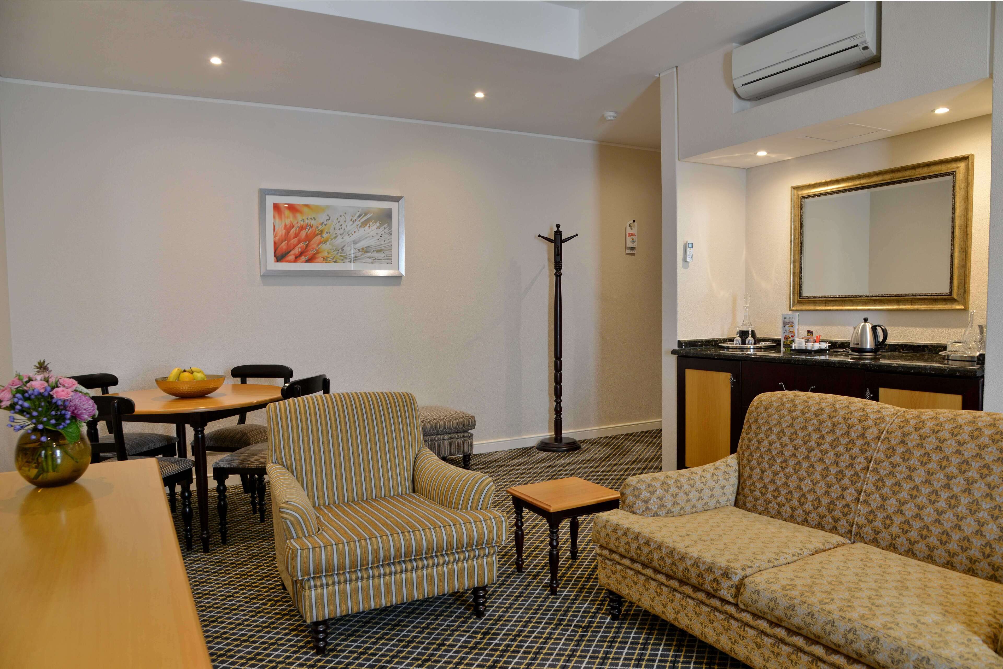 A hotel room with a dining area, a sofa, a patterned armchair, a small table, a floor lamp, a mirror, and a kitchen area with a coffee maker.