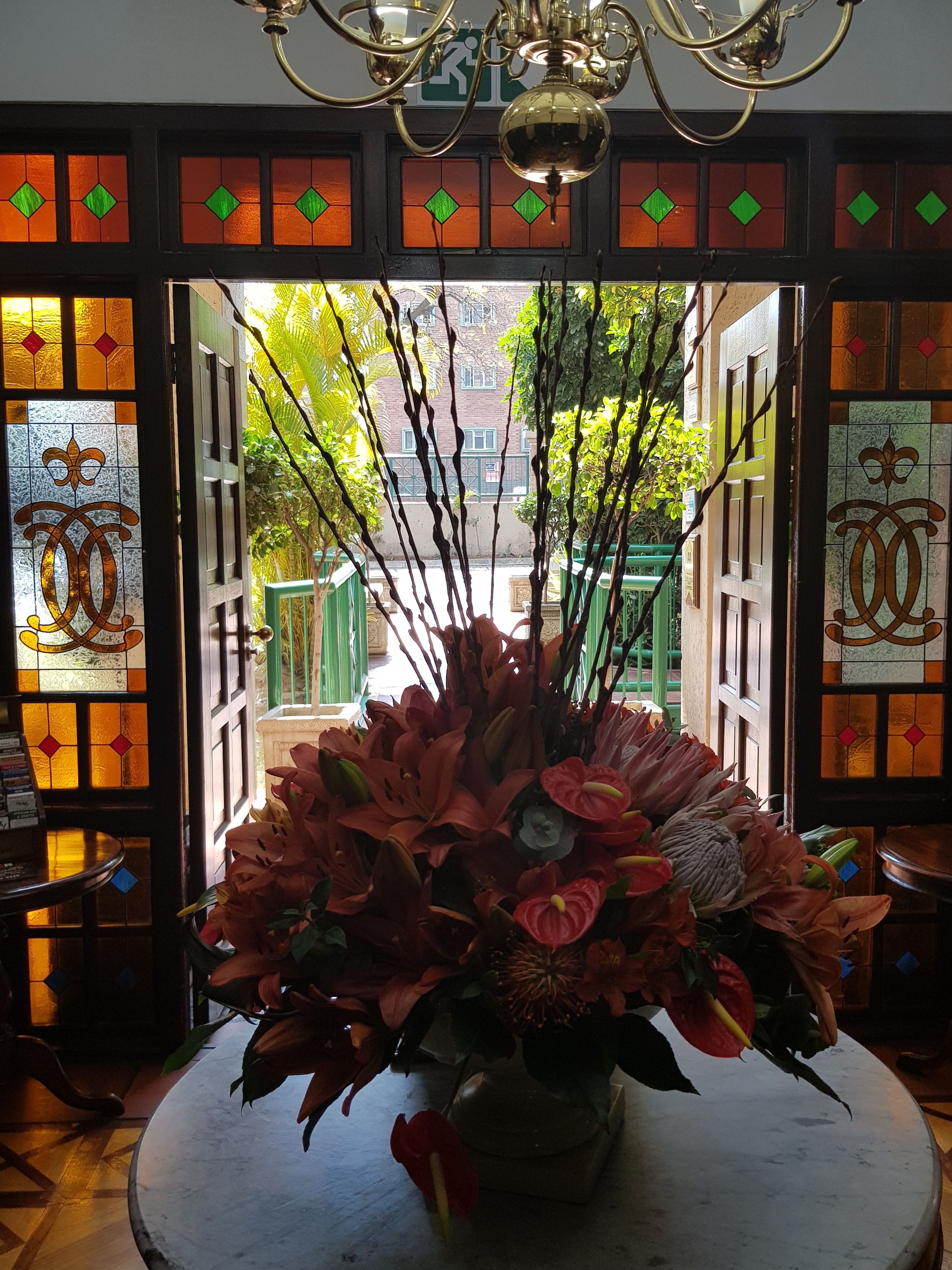 Una habitación con un gran arreglo floral sobre una mesa redonda, una araña y vistas de la calle a través de un arco con vidrieras.