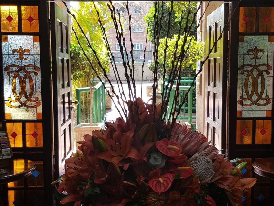 Una habitación con un gran arreglo floral sobre una mesa redonda, una araña y vistas de la calle a través de un arco con vidrieras.