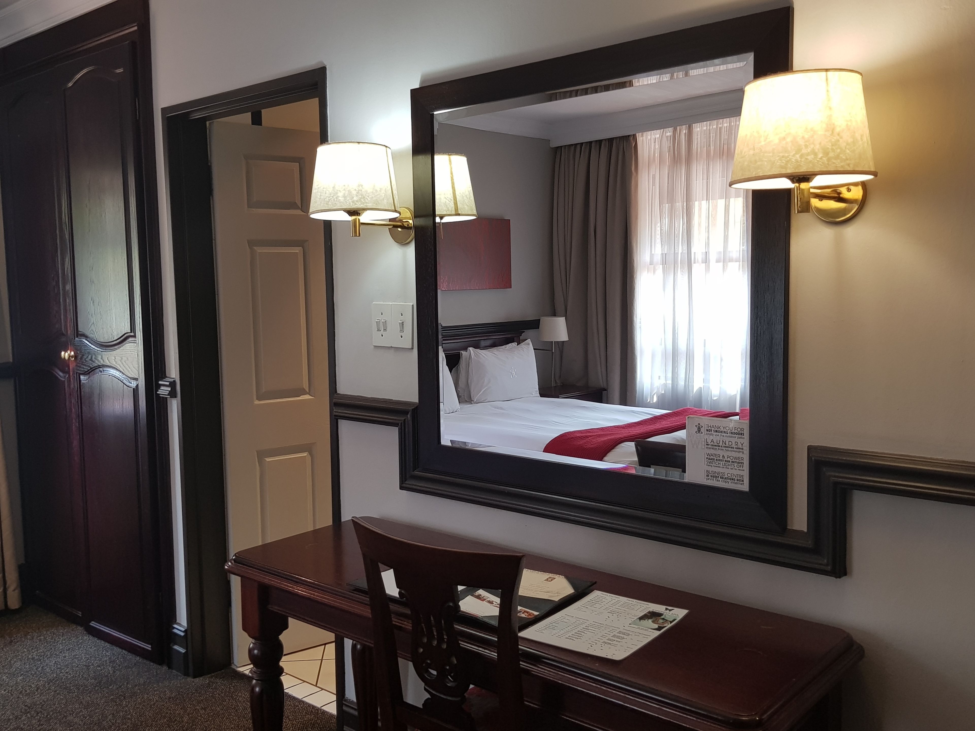 Habitación de hotel con cama, escritorio y espejo. Cuenta con dos lámparas de pared, una puerta y una ventana con cortinas.
