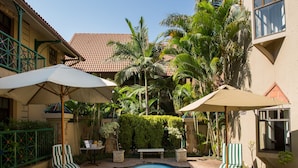 A swimming pool with lounge chairs and umbrellas, surrounded by palm trees and a building with a tiled roof.