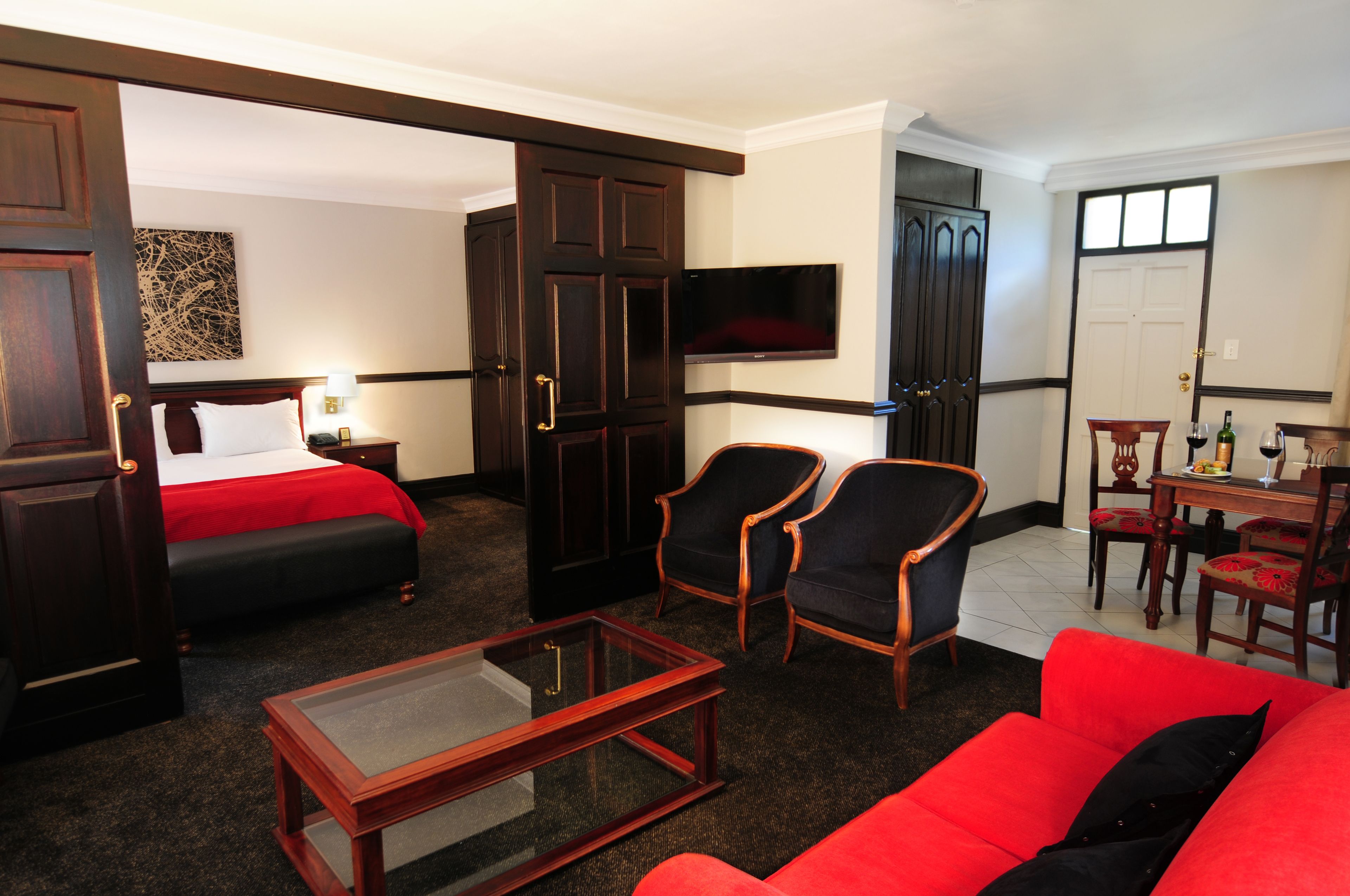Une chambre d’hôtel comprenant un lit, une télévision, deux fauteuils, un canapé rouge, une table basse et un coin repas avec des chaises et une table.