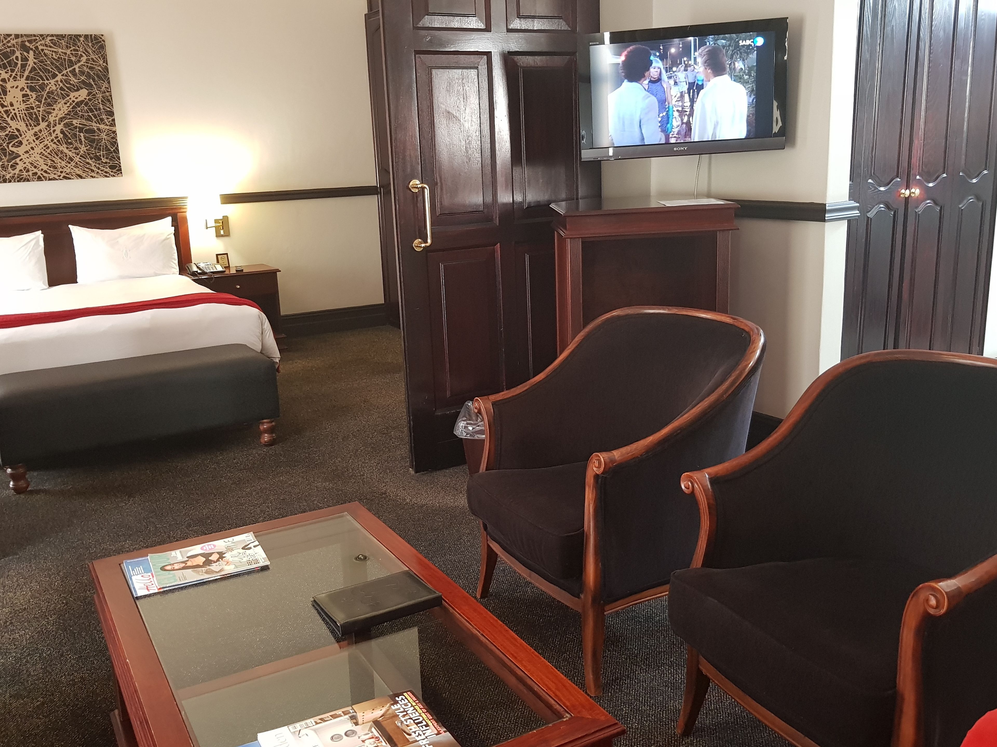 Una habitación de hotel con una cama, un televisor montado en la pared, dos sillones y una mesa de centro de vidrio.
