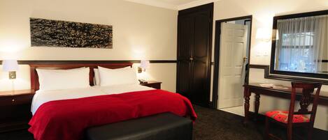 A hotel room with a large bed, a bench, a desk with a chair, a TV, and a door leading to a bathroom.