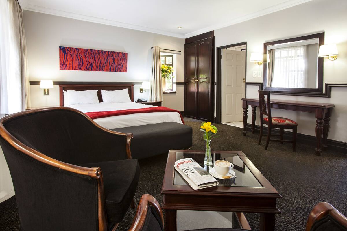 Ein Hotelzimmer mit Bett, Schreibtisch, Stuhl, Tisch mit Zeitung und Blumen in einer Vase.