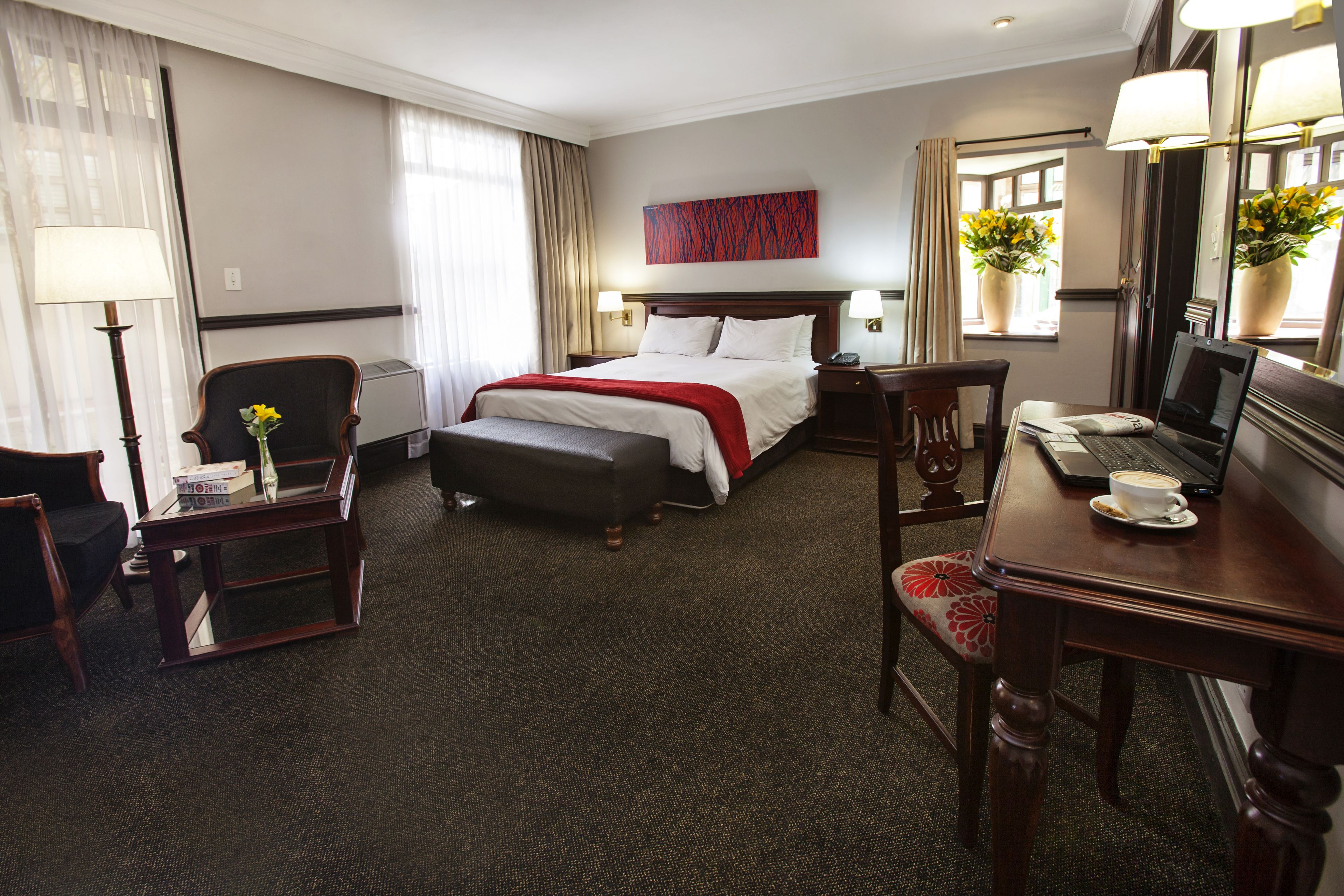 Une chambre d’hôtel avec un lit, un bureau avec un ordinateur portable, une chaise, une petite table avec un vase et une fenêtre avec des rideaux.
