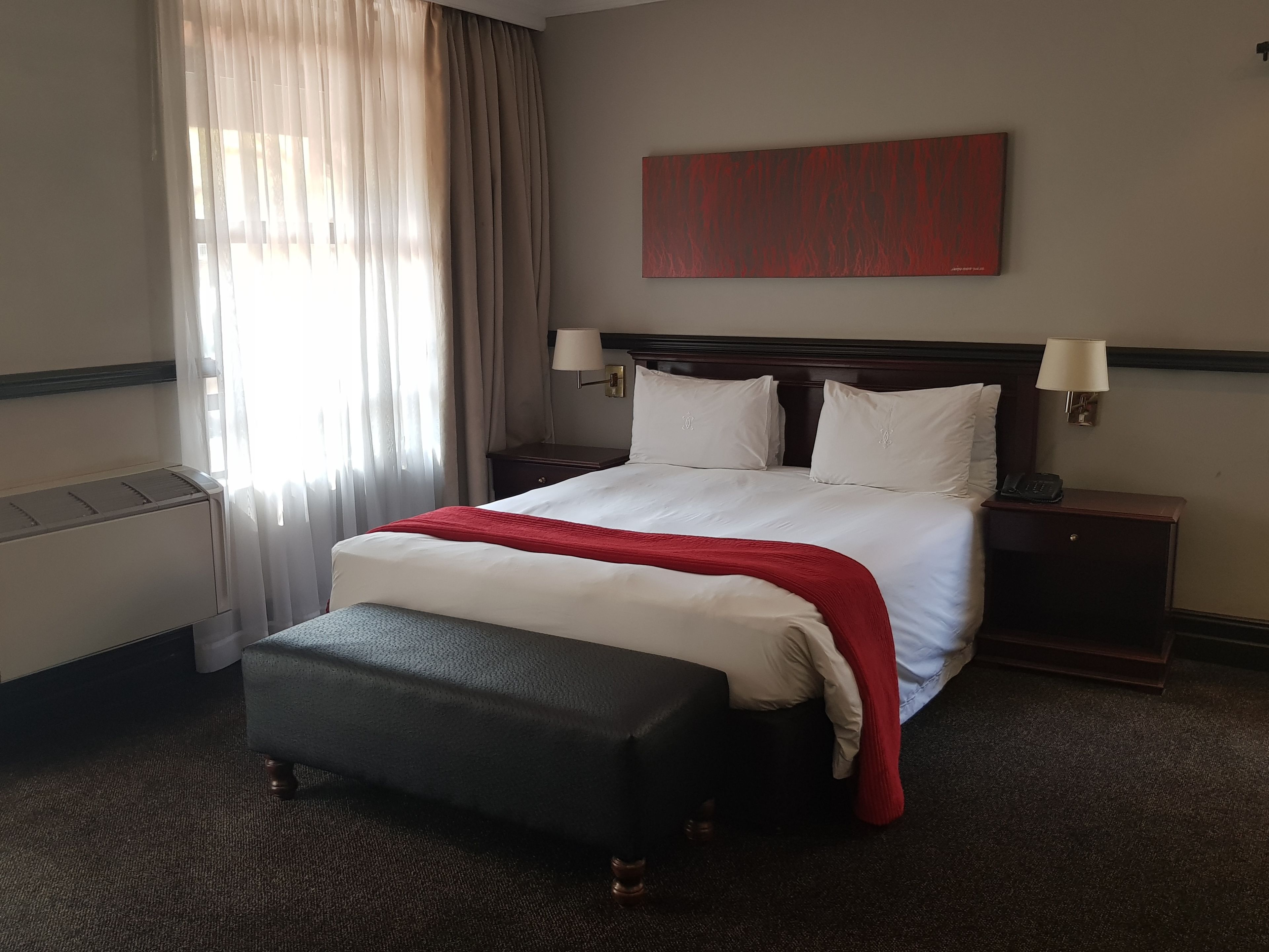 Una habitación de hotel con una cama grande, una mesita de noche con lámpara, una manta roja y una ventana con cortinas.