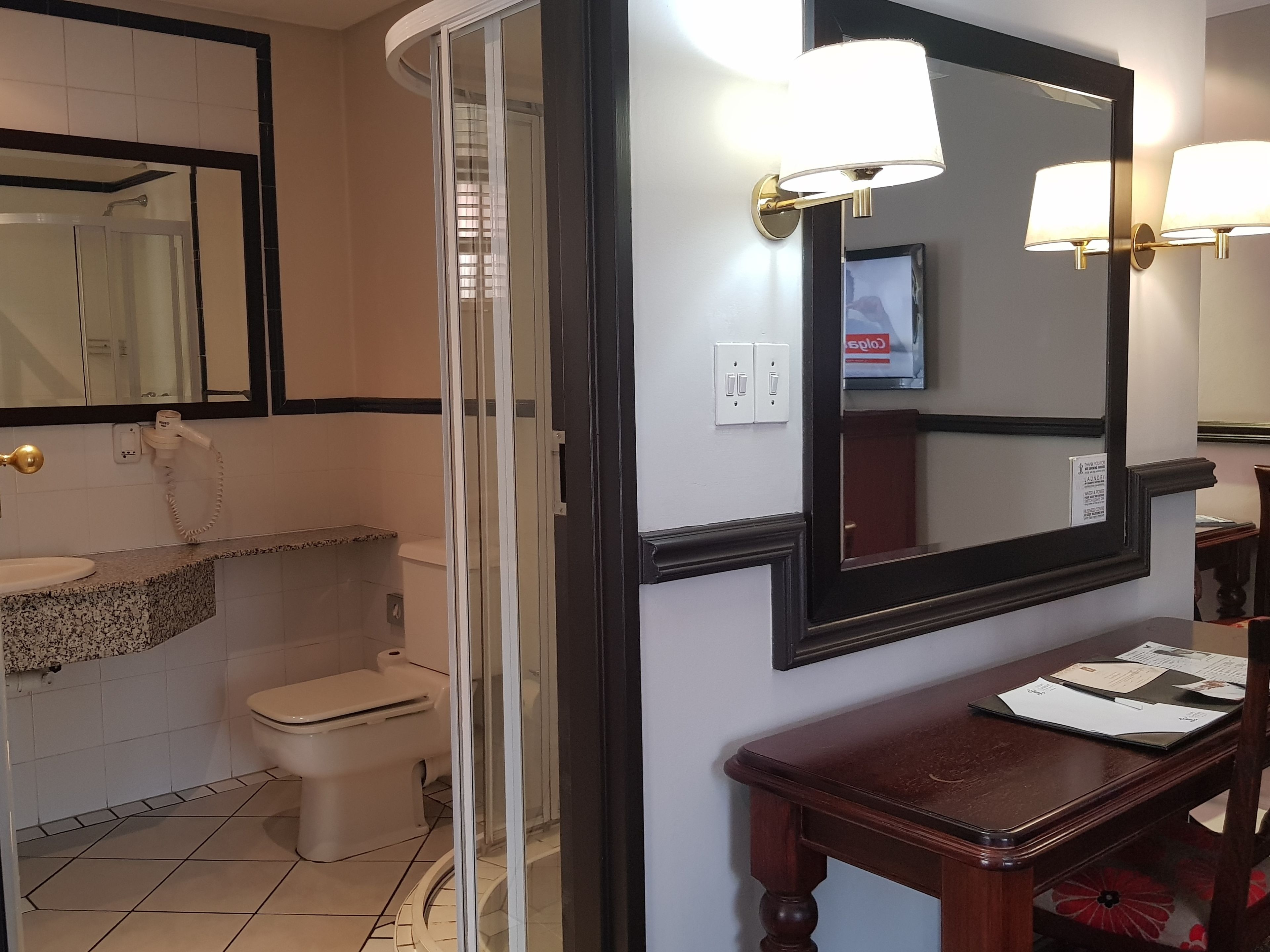 Un baño con inodoro, lavabo y ducha. Hay un espejo con un televisor encima, y una mesa de madera con papeles.