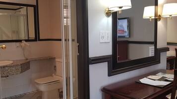 Un baño con inodoro, lavabo y ducha. Hay un espejo con un televisor encima, y una mesa de madera con papeles.