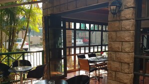 An outdoor cafe with tables and chairs, large windows, and a stone wall.