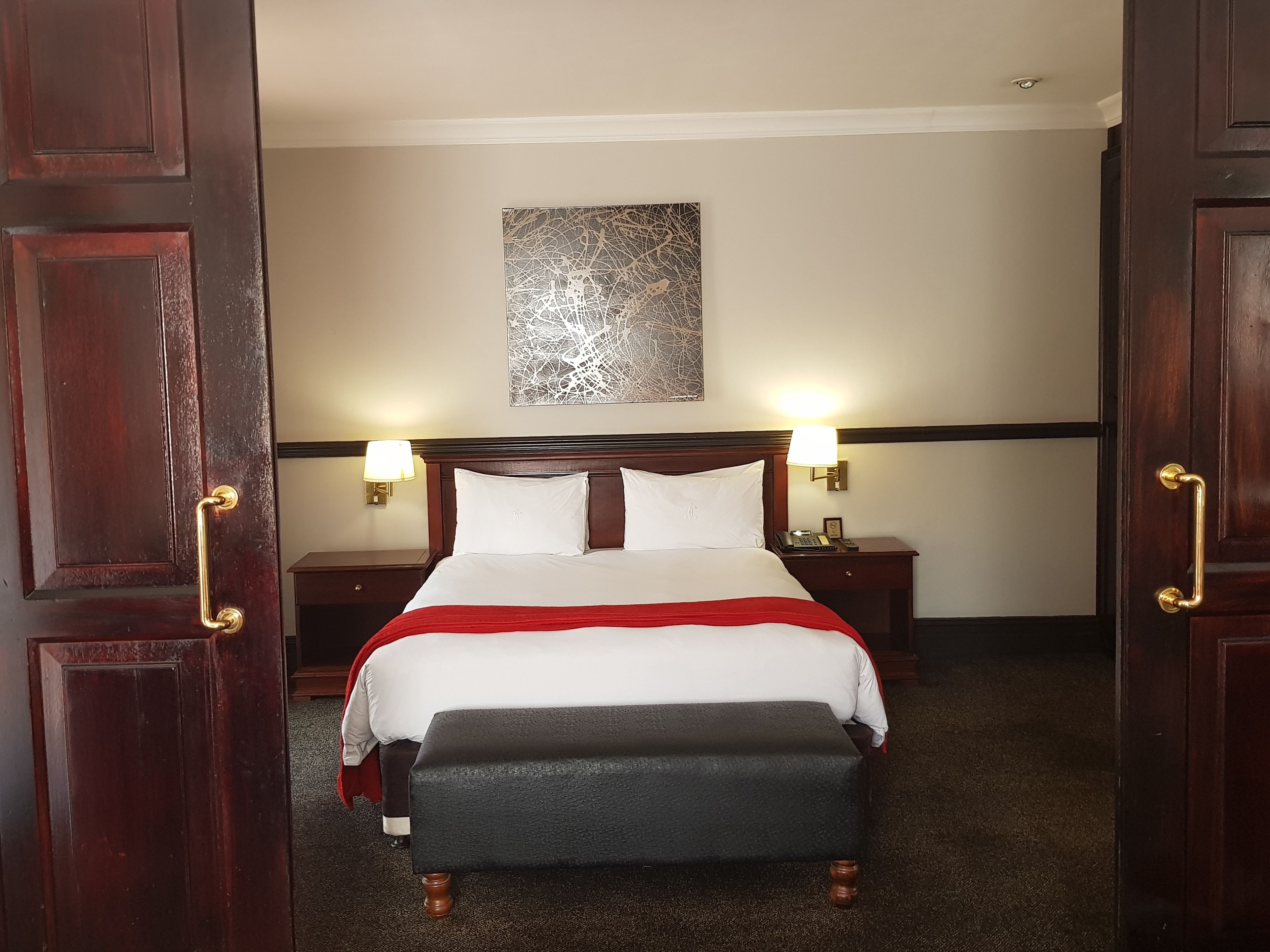 Una habitación de hotel con una cama grande, dos mesitas de noche, una lámpara de noche, un plaid rojo y una obra de arte abstracto colgada en la pared.