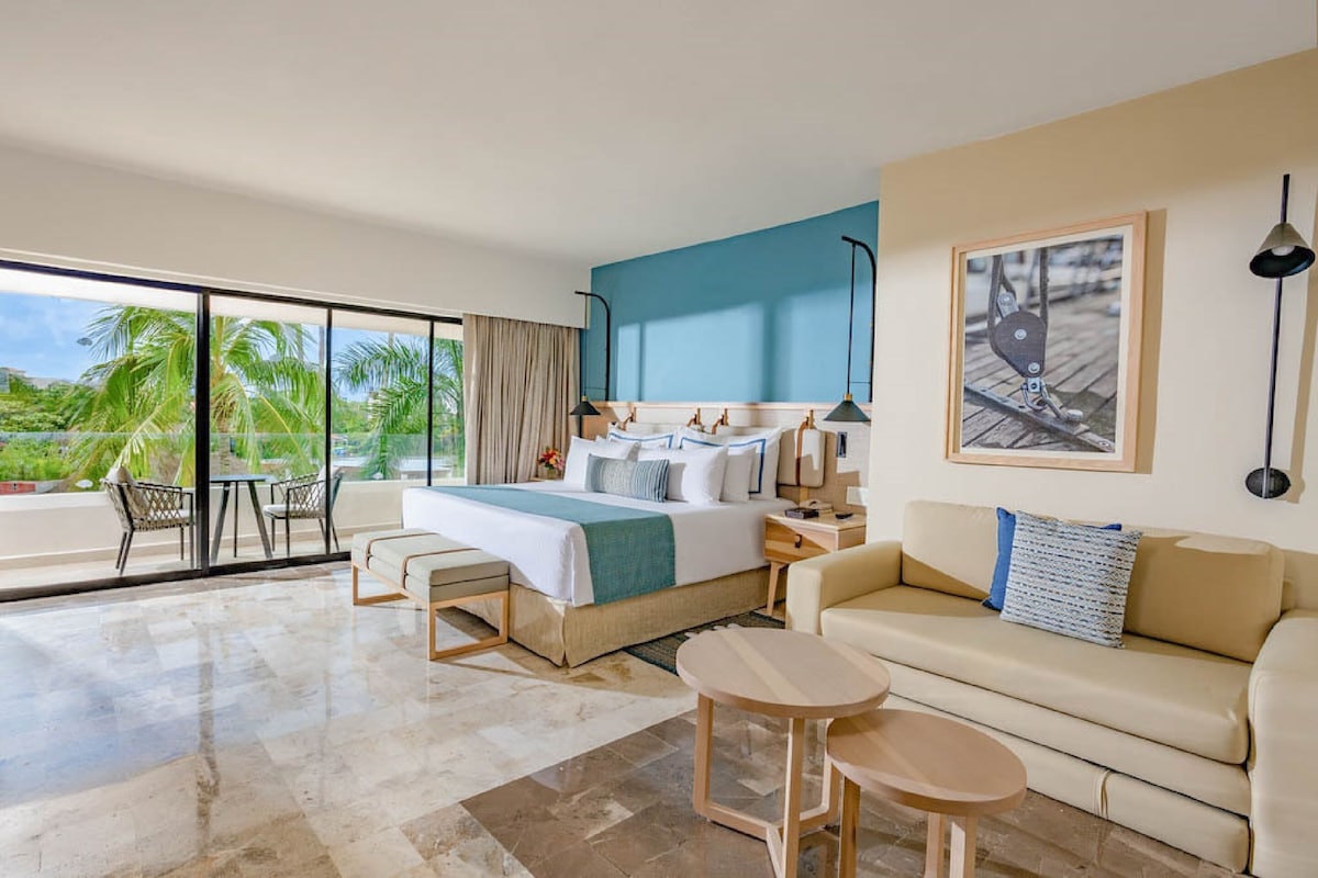 A modern hotel room with a large bed, a sofa, a small table, and a balcony with a view of palm trees. - Dreams Aventuras Riviera Maya - All Inclusive (Puerto Aventuras)