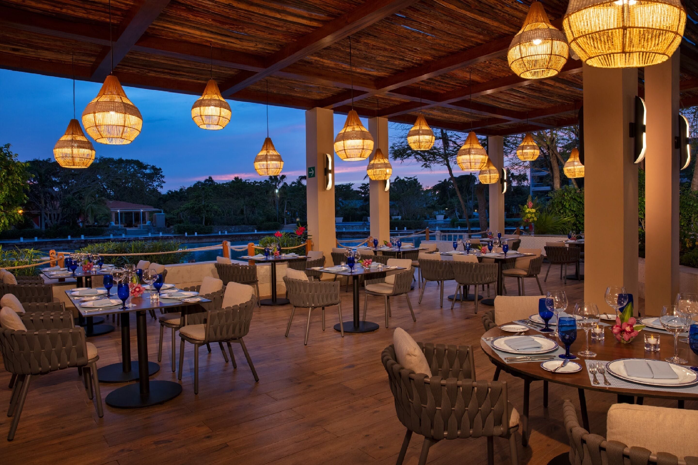 An outdoor dining area with tables set for guests, surrounded by columns and hanging lamps.